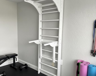 Könnte beinhalten: Eine weiße Kletterwand aus Holz mit gewölbtem Oberteil und mehreren Sprossen. Sie verfügt über eine Klimmzugstange und ein kleines, zusammenklappbares Regal. Die Wand ist an einer grauen Wand in einem Heim-Fitnessstudio montiert. Trainingsgeräte sind im Hintergrund zu sehen.