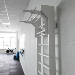 Puede incluir: Una espaldera de gimnasia blanca montada en la pared, que cuenta con una serie de pelda&ntilde;os horizontales y una extensi&oacute;n de barra de dominadas integrada en la parte superior. La estructura est&aacute; fijada verticalmente para ejercicios f&iacute;sicos.