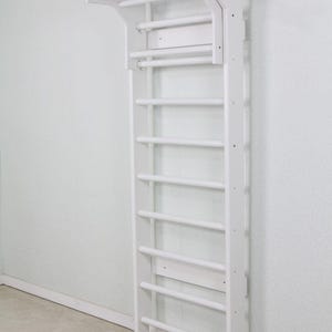 Puede incluir: Una espaldera de madera blanca montada en la pared con un dise&ntilde;o de escalera vertical, m&uacute;ltiples pelda&ntilde;os horizontales y una extensi&oacute;n de barra de dominadas integrada en la parte superior. Estructura de madera maciza con acabado blanco.