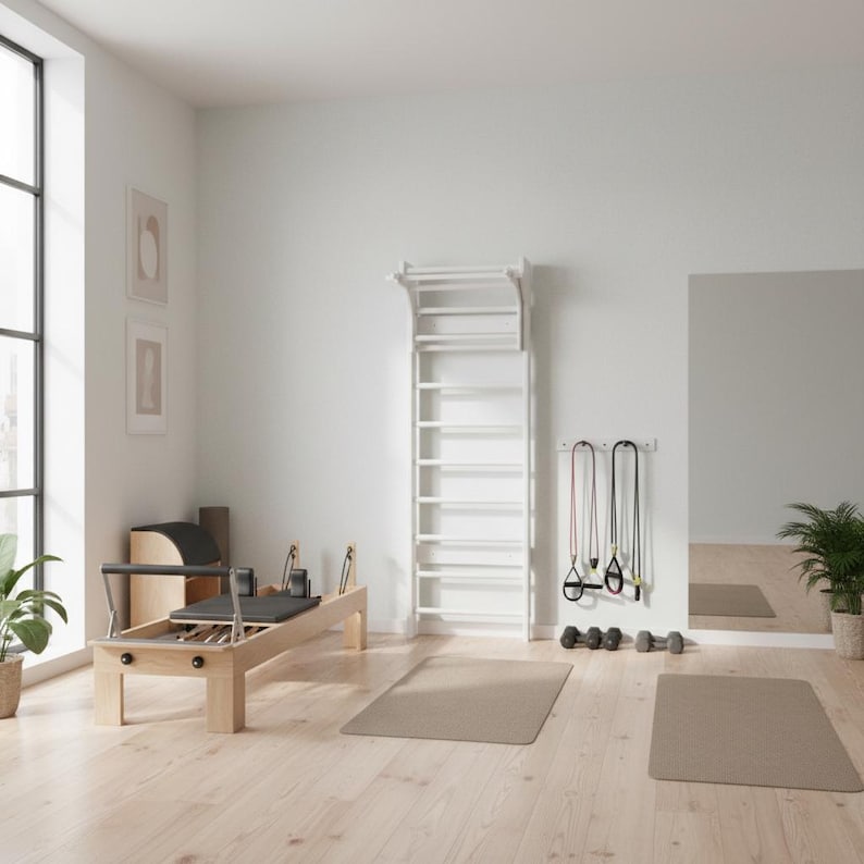 Puede incluir: Una espaldera de madera blanca montada en la pared con pelda&ntilde;os horizontales y una barra de dominadas superior, situada en una habitaci&oacute;n junto a una m&aacute;quina de Pilates de madera clara, dos bandas de resistencia y dos mancuernas peque&ntilde;as.