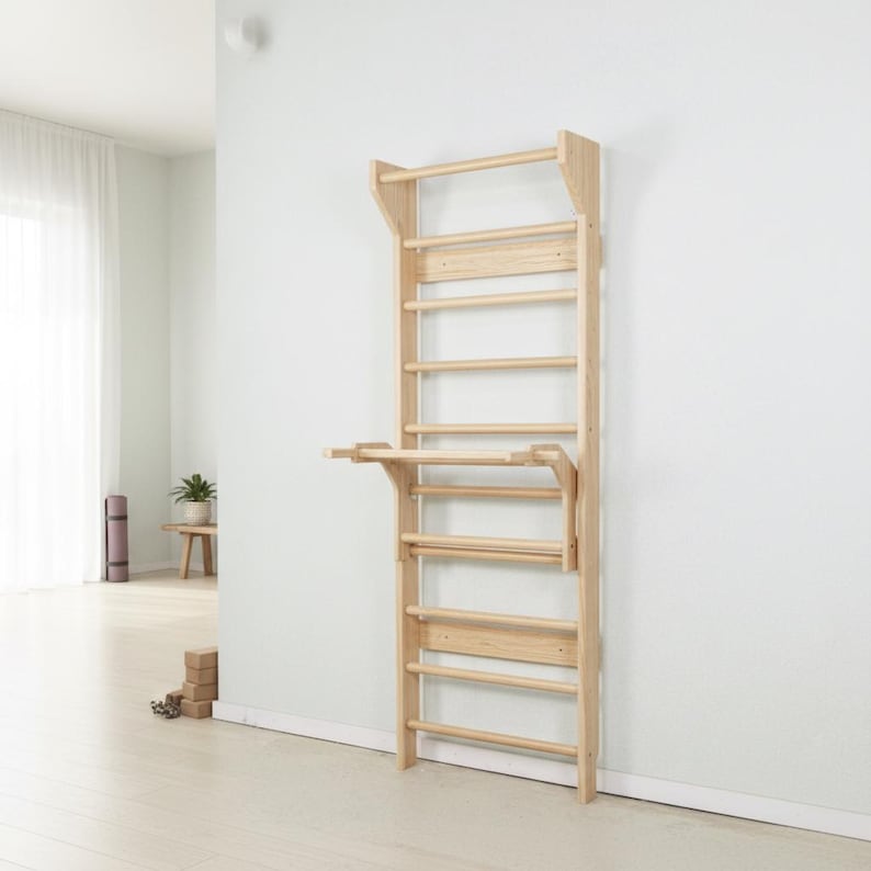 Puede incluir: Una unidad de espaldera de madera de color claro montada en la pared que cuenta con una serie de pelda&ntilde;os horizontales y una plataforma horizontal plegable unida a la secci&oacute;n central, dise&ntilde;ada para ejercicios f&iacute;sicos y escalada.