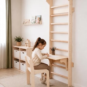 May include: A wooden climbing frame with a desk and chair attached. A child is drawing at the desk. A shelf with books and a toy house are visible. The room has a neutral colour scheme with a rug and curtains.