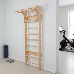 May include: A wooden wall-mounted Swedish ladder with multiple horizontal rungs for climbing and exercise. The ladder is made of light-colored wood and is attached to a white wall. A blue exercise mat and dumbbells are on the floor.
