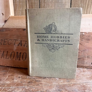 Puede incluir: Un libro antiguo titulado "HOME HOBBIES & HANDICRAFTS" con una cubierta verde claro. La cubierta presenta una ilustración negra de una cesta, un jarrón y otros objetos. El libro está sobre una superficie de madera.