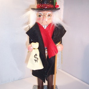 Op de afbeelding: Een houten notenkrakerfiguur van een vooraanstaande heer in een zwarte hoge hoed met glitter, een rode sjaal en een zwarte jas. Hij houdt een geldbuidel met een dollarteken en een wandelstok vast. De figuur staat op een zilveren bakstenen voet.