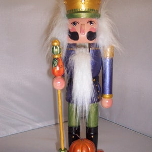 May include: A wooden nutcracker figurine with a purple and green outfit, a white beard, and a gold crown. He is holding a staff with a green and orange pumpkin on top. The nutcracker is standing on a base with a pumpkin and fall leaves.