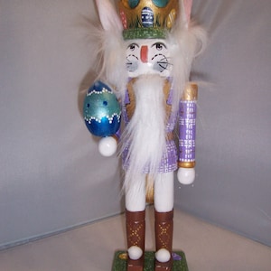 May include: A white bunny nutcracker with a purple and gold crown, holding a blue and green Easter egg. The nutcracker is wearing brown boots and standing on a green base with Easter eggs.