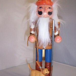 May include: A wooden nutcracker in the form of a hunter, wearing an orange hat and brown jacket, holding a rifle and a leash attached to a small dog. The nutcracker has a white beard and is standing on a green base.