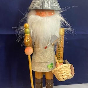 May include: A wooden gnome figurine with a long white beard, wearing a gray hat and apron. The gnome holds a walking stick, a basket, and a small shovel. The base features mushrooms and autumn leaves.