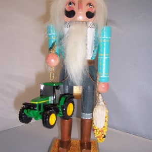 Peut inclure: Une figurine de casse-noisette en bois représentant un fermier portant un chapeau de paille, une chemise à carreaux bleus et blancs et un pantalon marron. Il tient un petit tracteur jouet vert et un sac de maïs. Le casse-noisette est debout sur une base avec de l'herbe verte et du foin jaune.