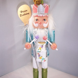 May include: A teal and white nutcracker with a pink crown, holding drumsticks and a balloon that says "Happy Birthday".