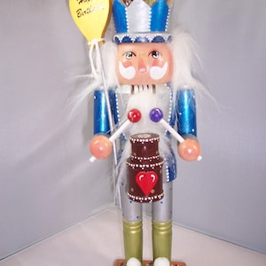 May include: A blue and silver nutcracker with a white beard and mustache, wearing a crown and holding a chocolate cake with a red heart. The nutcracker is holding drumsticks and a balloon that says "Happy Birthday".
