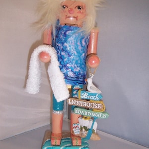 May include: A wooden nutcracker doll wearing a blue and white patterned dress, a white towel, and a straw hat. The doll is holding a small white bird and standing on a base with a sign that reads "Beach Lighthouse Boardwalk".