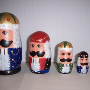 May include: A set of six wooden nesting dolls, each painted to resemble a nutcracker soldier. The dolls vary in size, with detailed facial features, mustaches, and ornate hats. The color palette includes blue, red, green, gold, and white.