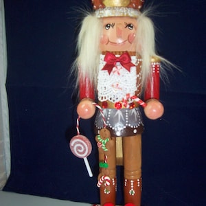 May include: A wooden nutcracker doll with a crown, red and white accents, and a lollipop. The doll has a red bow tie, a white lace vest, and a brown skirt. The base is red with a decorative pattern. A gingerbread man and candy canes are attached to the legs.
