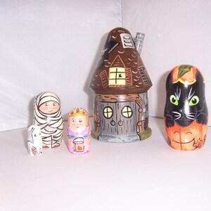 Pode incluir: Um conjunto de cinco bonecas russas com tema de Halloween. A boneca maior é uma casa marrom com uma porta e janelas pretas. As outras bonecas são uma múmia, uma princesa, um gato preto e um fantasma branco.
