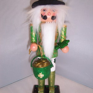 May include: A green and gold nutcracker dressed as a leprechaun with a white beard, black hat, and a pot of gold. He is holding a shamrock in his right hand.