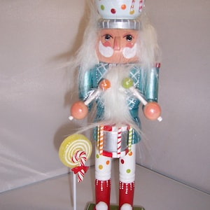 Könnte beinhalten: Ein skurriler Zuckerstangen-Nussknacker mit weißem Bart, blau-grünem gemustertem Mantel und weißer Hose mit roten und gelben Punkten. Der Nussknacker hält Zuckerstangen und trägt eine Schüssel Süßigkeiten auf dem Kopf.
