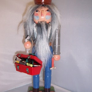 May include: A wooden nutcracker figurine dressed as a construction worker. He is wearing a brown hard hat, a blue shirt, and blue jeans. He is holding a red toolbox with tools inside. The nutcracker is standing on a brick base.