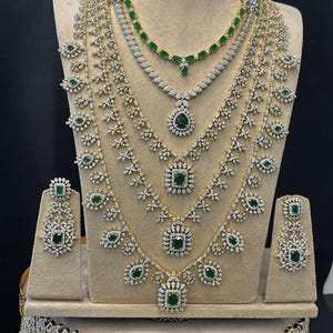 Może przedstawiać: Zestaw biżuterii składający się z kilku naszyjników, kolczyków i bransoletki. Elementy w kolorze złota zdobione są przezroczystymi i szmaragdowozielonymi kamieniami. Zestaw jest prezentowany na beżowym manekinie.