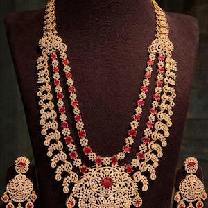 May include: A three-strand gold-toned necklace with a large pendant and matching earrings. The necklace and earrings are adorned with clear stones and red gemstones. The jewelry is displayed on a dark brown bust.
