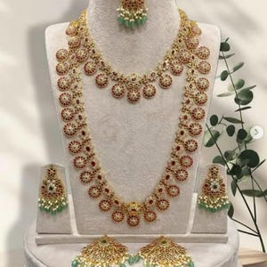 May include: A gold-toned jewelry set featuring a necklace, earrings, and a headpiece. The necklace and earrings have a circular design with red and white accents. The headpiece and earrings have green and white accents. The jewelry is displayed on a mannequin.