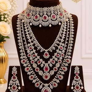 May include: A luxurious jewelry set featuring a choker, layered necklaces, and matching earrings. The set is adorned with sparkling white stones and vibrant red gemstones. The jewelry is displayed on a brown bust, with the brand name "sstraditions" visible.