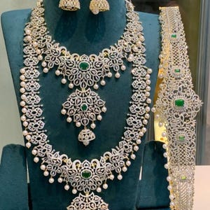 Könnte beinhalten: Ein schillerndes Schmuckset mit Halskette, Ohrringen und Armband. Das Set ist mit klaren Steinen, Kunstperlen und grünen Akzenten verziert. Halskette und Armband haben aufwendige Blumenmuster. Die Ohrringe sind glockenförmig mit einem Perltropfen.