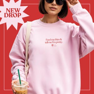 May include: A light pink sweatshirt with the text "Feed me fries & tell me I'm pretty" in red. The sweatshirt has a cupcake and smiley face graphic. The words "NEW DROP" are in a starburst shape in the upper left corner.