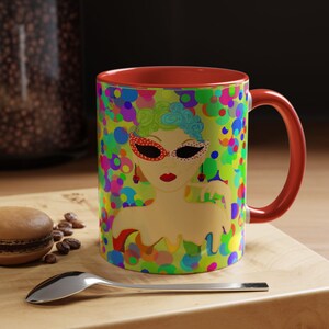 May include: A ceramic coffee mug with a red interior and handle. The exterior features a colorful illustration of a woman wearing a mask, surrounded by a pattern of colorful circles. The mug sits on a wooden surface.