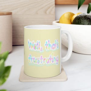 May include: A light yellow ceramic mug with the text "Well, that's frustrating" in pastel colours. The mug sits on a square beige coaster. The mug has a white handle and is in a kitchen setting.