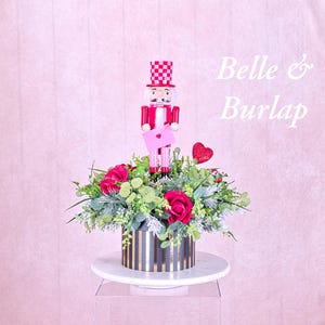 May include: A decorative floral arrangement featuring a red and white nutcracker holding a pink envelope, surrounded by red roses and greenery. The arrangement sits in a black and gold striped container on a white pedestal. The text "Belle & Burlap" is visible.