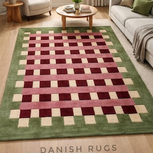 Puede incluir: Una alfombra rectangular con un borde verde y un patrón tejido de cuadrados burdeos, crema y rosas. La alfombra está colocada sobre un suelo de madera en un salón. El texto "DANISH RUGS" es visible.