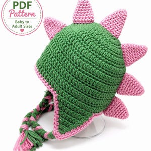 May include: A green crocheted hat with pink spikes along the top and back, featuring braided green and pink earflaps with tassels. A circular badge in the upper left corner contains the text PDF Pattern Baby to Adult Sizes with a heart icon.