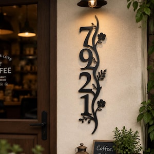 May include: A vertical black metal house number sign displaying the digits 7, 9, 2, and 1, integrated with a decorative vine motif featuring leaves and small flowers. The sign is mounted on a light-colored wall next to a dark brown door.