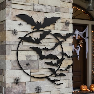 May include: Black metal bat wall decoration featuring a circular design, surrounded by smaller bats, spiders, and spiderwebs. The Halloween-themed decor is mounted on a stone wall, with a decorated porch in the background.