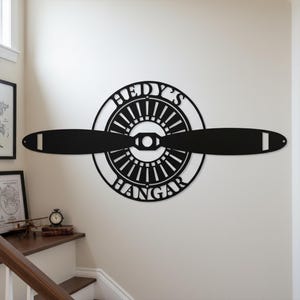 May include: Black metal wall art featuring an airplane propeller design. The circular center displays the text "HEDY'S HANGAR" in a bold, stylized font. The propeller blades extend horizontally, creating a striking aviation-themed decor piece.