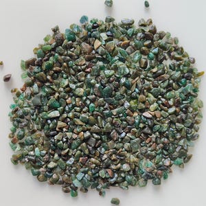 May include: A pile of small, irregularly shaped green and brown stones. The stones vary in shade, from light green to dark brown, and are scattered across a white surface. The stones appear to be polished or tumbled.