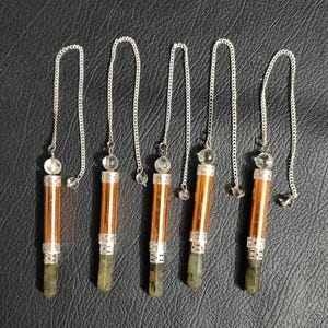 May include: Five pendulums with silver chains, clear crystal beads, and copper-coloured cylinders. Each pendulum has a faceted, green-grey crystal point at the base. The pendulums are arranged on a black surface.