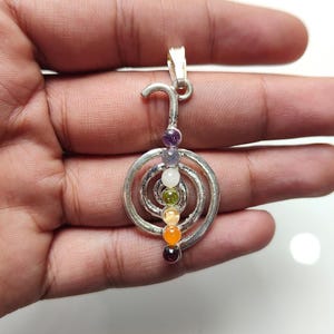 May include: Silver pendant with a spiral design and seven coloured gemstone beads. The beads are arranged vertically, displaying a range of colours from purple to red. The pendant has a curved top for a chain.