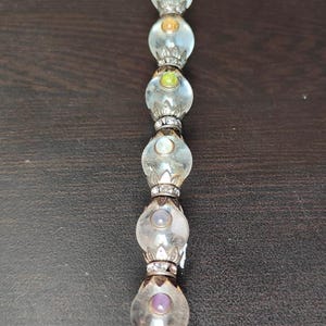 May include: A clear glass wand with a series of round, colourful beads. The beads are accented with silver-coloured metal and contain small, coloured gems. The wand is a decorative item.