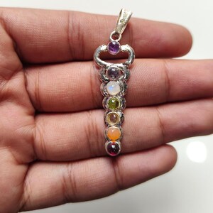 May include: A silver pendant with a key-shaped design, adorned with a sequence of seven gemstones. The gemstones are in a range of colours, including purple, blue, green, and orange. The pendant is designed to be worn as jewellery.