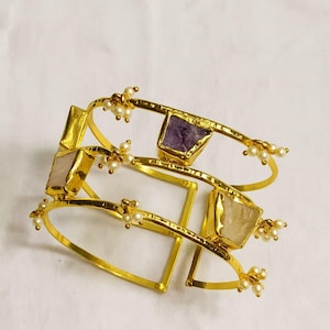 Peut inclure: Bracelets manchettes dorés avec des accents de pierres précieuses rectangulaires violettes, roses et crème. Les bracelets comportent de délicats groupes de perles. Bijoux pour le poignet, sur fond blanc.