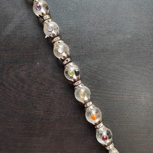 May include: A clear crystal wand with a silver-coloured metal handle and accents. The wand features a series of clear, spherical glass beads, each containing a different coloured gemstone. The wand is approximately 25 cm long.