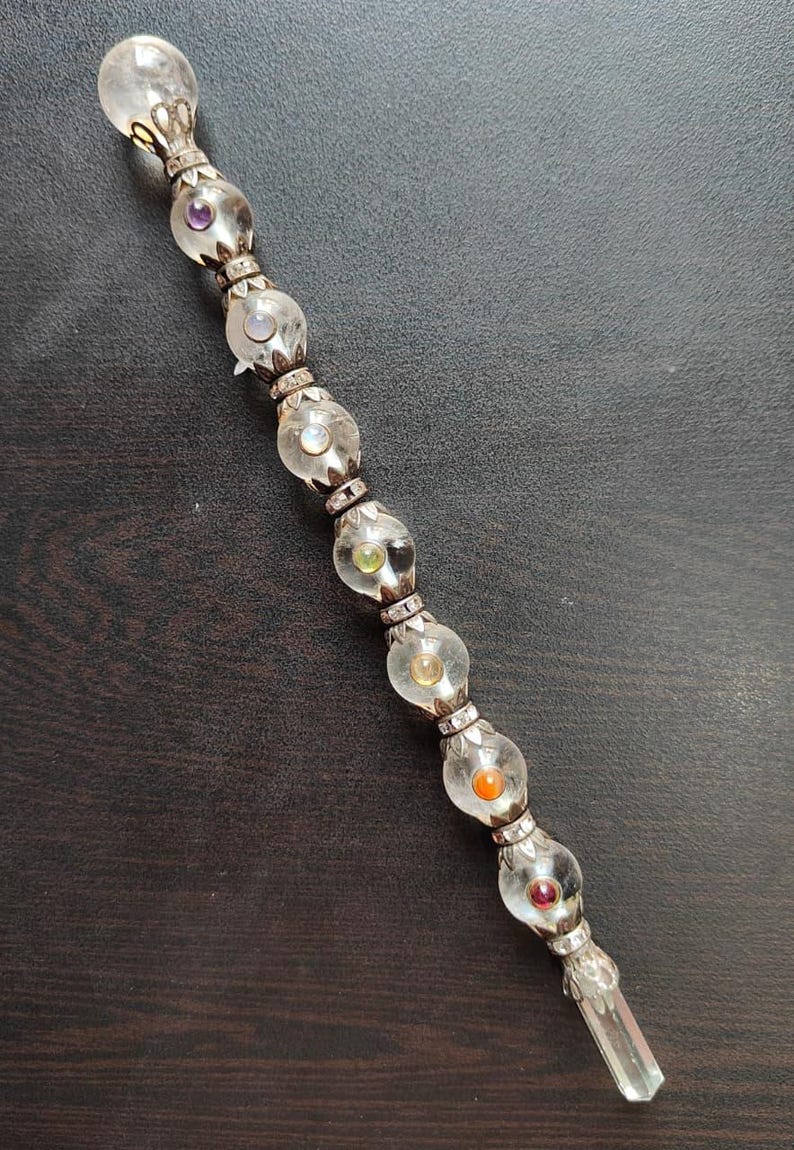 May include: A clear crystal wand with a silver-coloured metal handle and accents. The wand features a series of clear, spherical glass beads, each containing a different coloured gemstone. The wand is approximately 25 cm long.