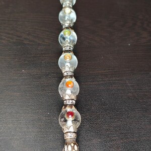 May include: A decorative crystal wand with clear glass orbs and colourful gemstones. The wand features a clear, faceted crystal at the base and is accented with silver-coloured metal. The orbs contain a variety of colours, including red, orange, green, and purple.