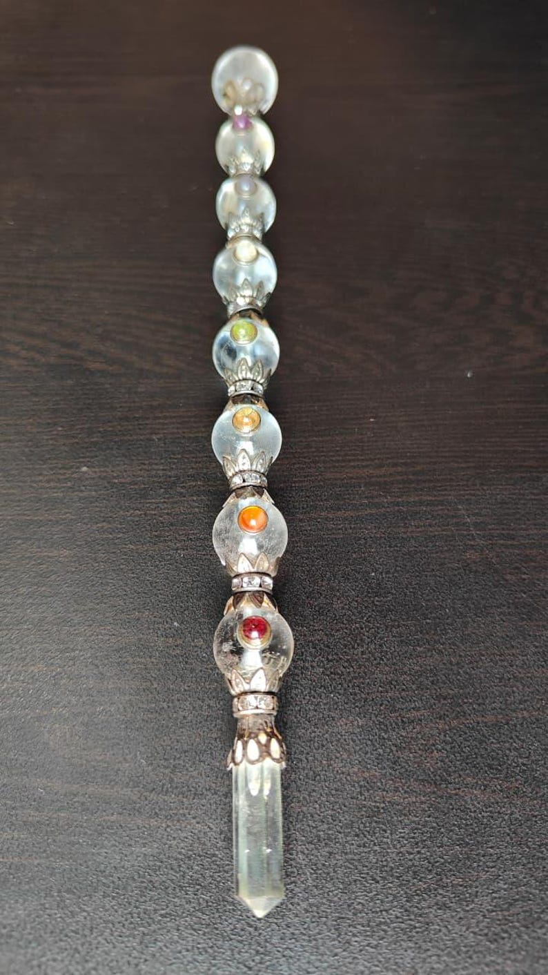 May include: A decorative crystal wand with clear glass orbs and colourful gemstones. The wand features a clear, faceted crystal at the base and is accented with silver-coloured metal. The orbs contain a variety of colours, including red, orange, green, and purple.