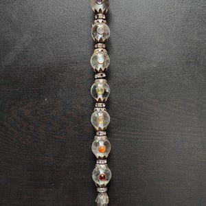 May include: A clear glass wand with a rounded top and a faceted crystal at the base. The wand is adorned with clear glass orbs and colourful gemstones, including purple, yellow, and orange. The wand has silver-coloured metal accents.
