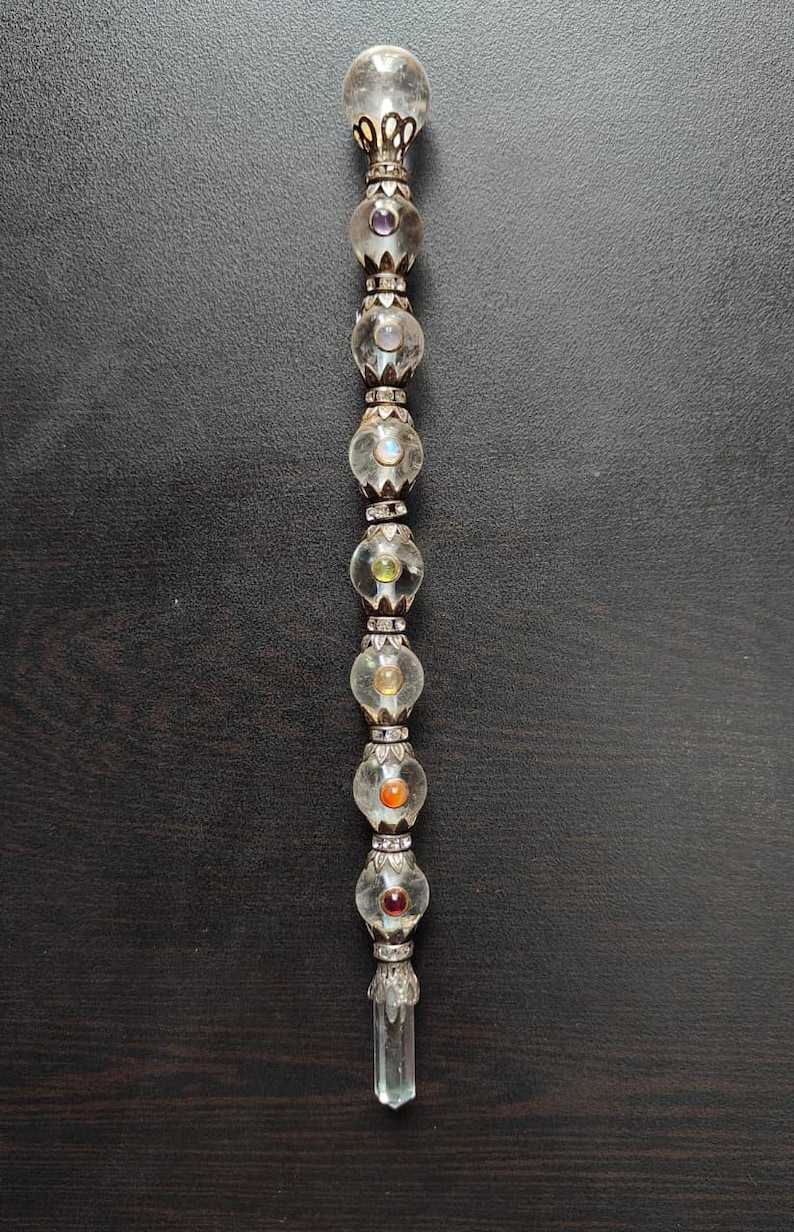 May include: A clear glass wand with a rounded top and a faceted crystal at the base. The wand is adorned with clear glass orbs and colourful gemstones, including purple, yellow, and orange. The wand has silver-coloured metal accents.