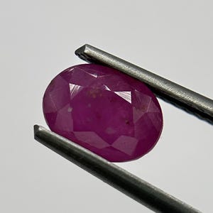 Può includere: Una gemma di rubino ovale e sfaccettata tenuta da una pinzetta di metallo. Il rubino è di un colore rosso intenso e saturo, con inclusioni interne visibili. La pinzetta è color argento e lo sfondo è bianco.